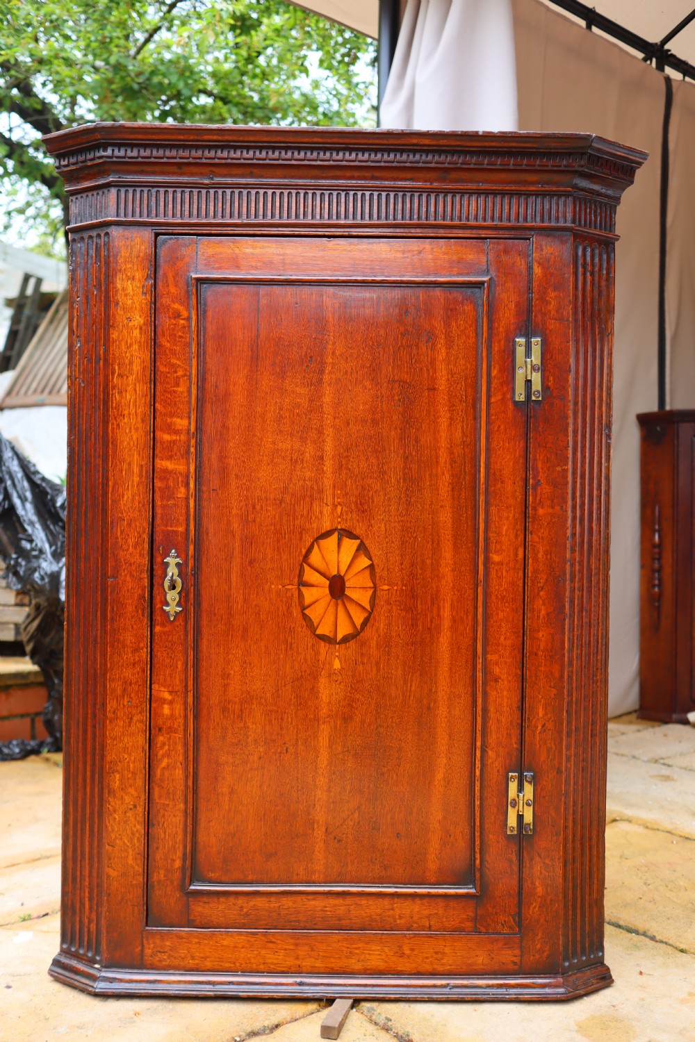 An antique oak corner cupboard featuring a single door with central oval starburst inlay, fluted pilasters, brass H-hinges, and a dentil moulding cornice. View VIII