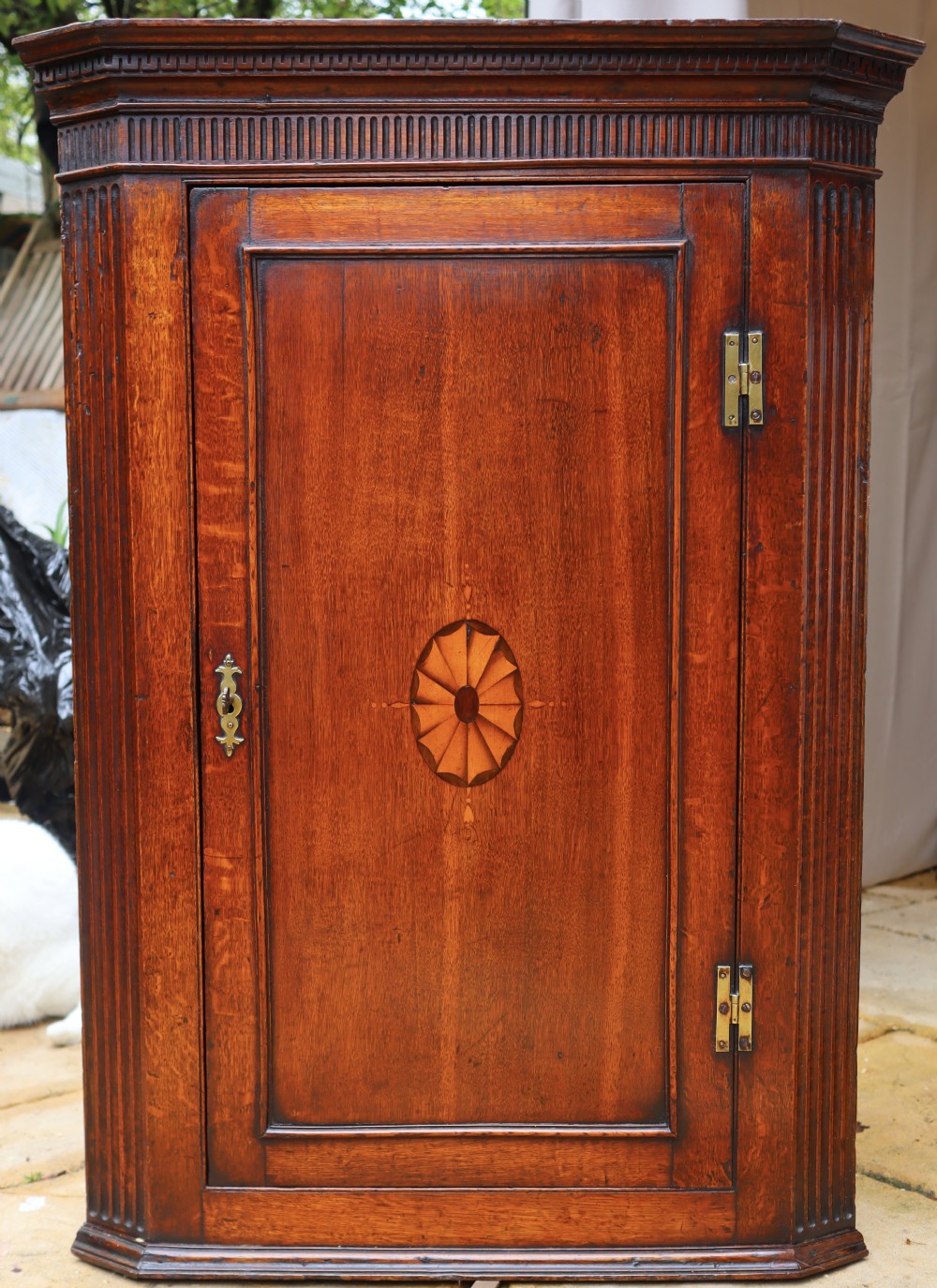 An antique oak corner cupboard featuring a single door with central oval starburst inlay, fluted pilasters, brass H-hinges, and a dentil moulding cornice.