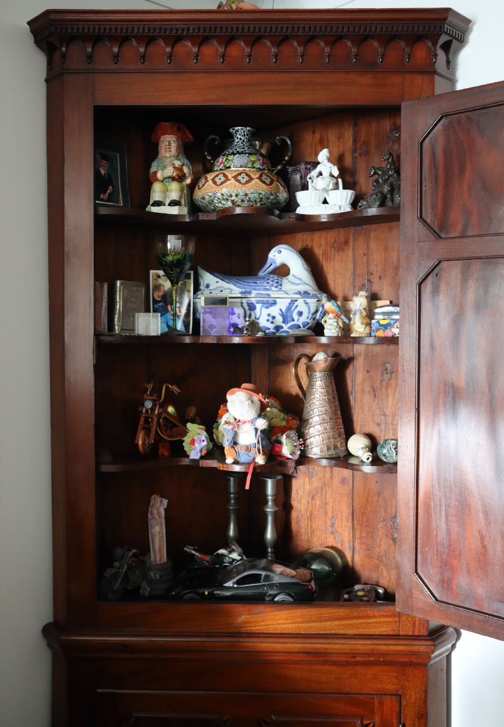 A tall, free-standing antique mahogany corner cupboard featuring two stacked panel doors decorated with carved corner details and top dentil moulding. View VIII