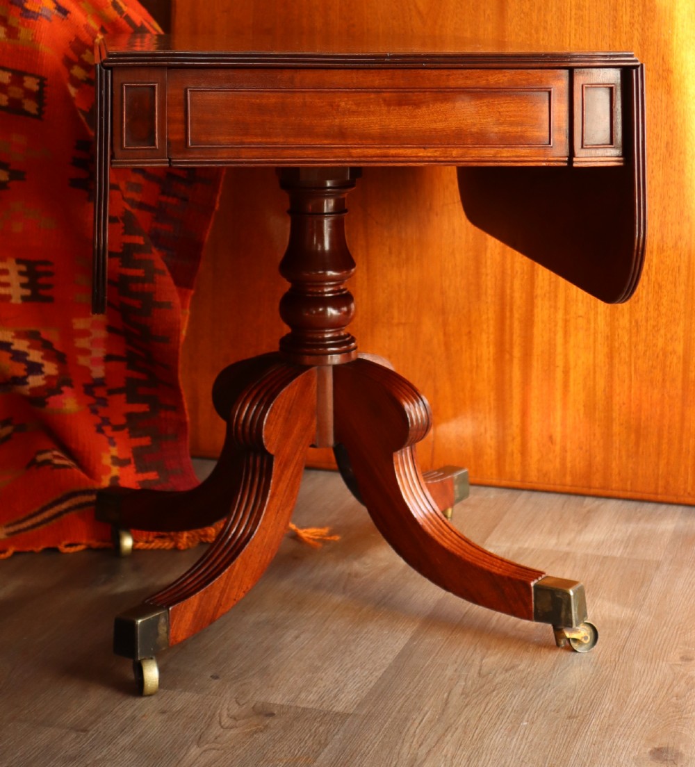 A mahogany pedestal dining table featuring drop-leaf sides, turned central column, four reeded splayed legs, and brass castors.
