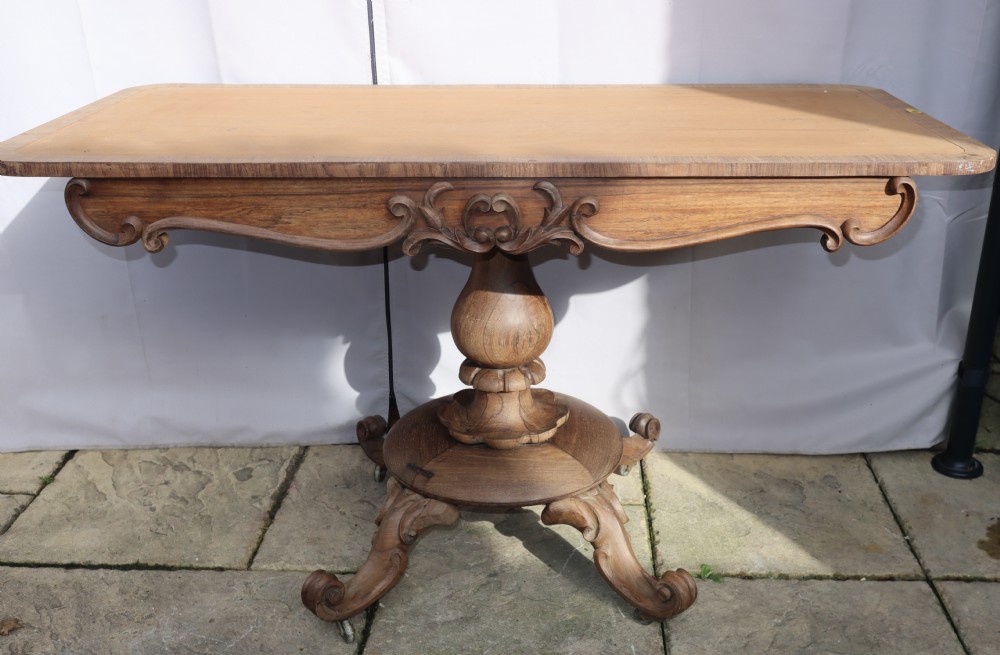 A carved rosewood centre table with an inset top, carved scroll apron, central baluster pedestal, and carved scroll feet on castors.