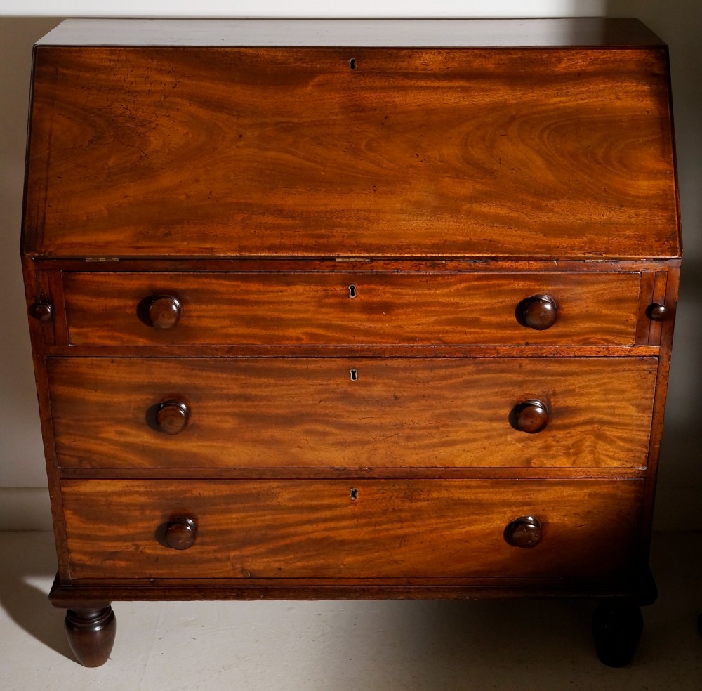 An antique warm brown mahogany bureau featuring a slant-front lid above three long drawers with turned wooden handles, set on turned feet. View III