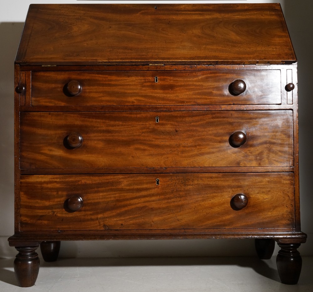 An antique warm brown mahogany bureau featuring a slant-front lid above three long drawers with turned wooden handles, set on turned feet.
