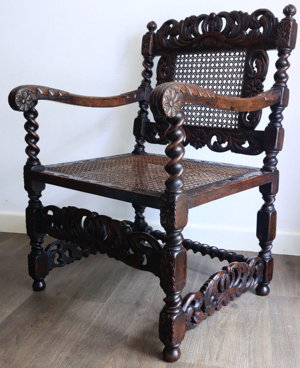 A dark carved oak armchair featuring barley twist supports, a woven cane back panel, and intricate foliate crown carving on the cresting rail. View IV