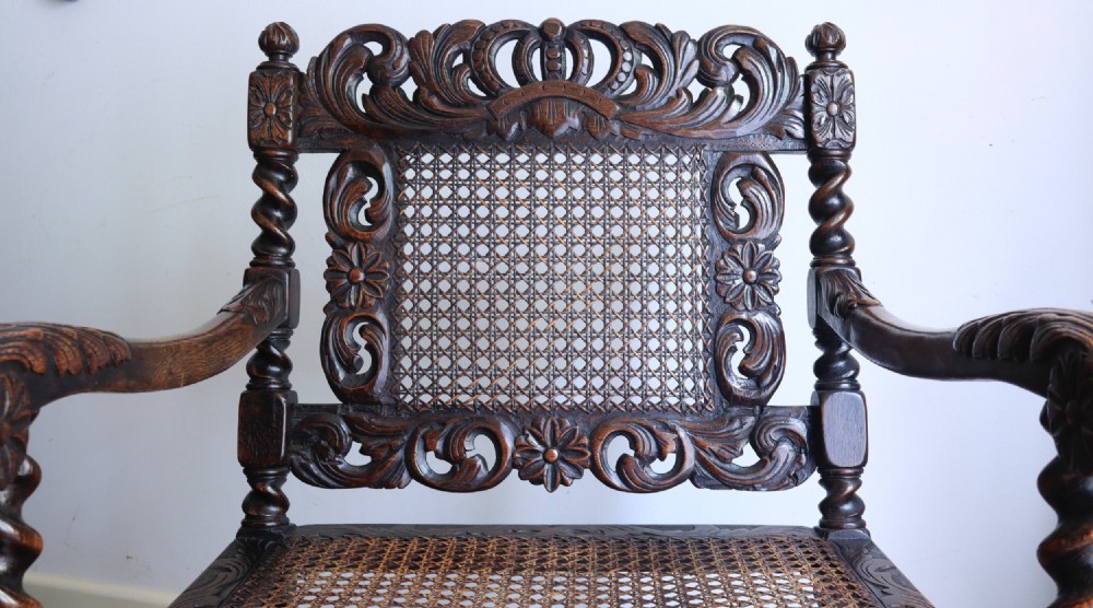 A dark carved oak armchair featuring barley twist supports, a woven cane back panel, and intricate foliate crown carving on the cresting rail. View V