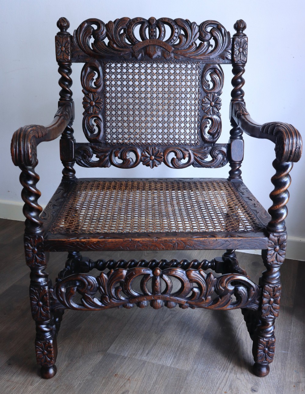 A dark carved oak armchair featuring barley twist supports, a woven cane back panel, and intricate foliate crown carving on the cresting rail. View VI
