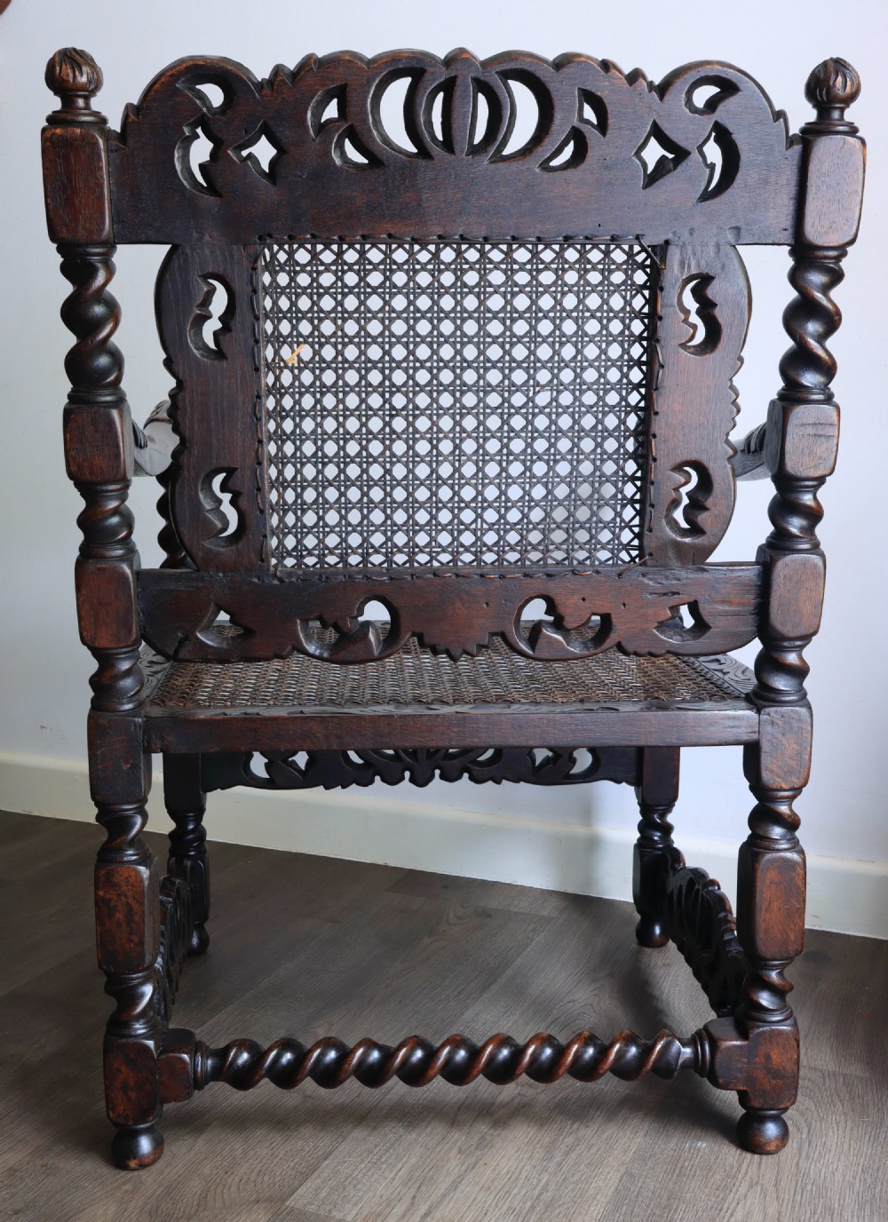 A dark carved oak armchair featuring barley twist supports, a woven cane back panel, and intricate foliate crown carving on the cresting rail. View VII