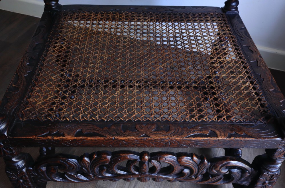A dark carved oak armchair featuring barley twist supports, a woven cane back panel, and intricate foliate crown carving on the cresting rail. View IX