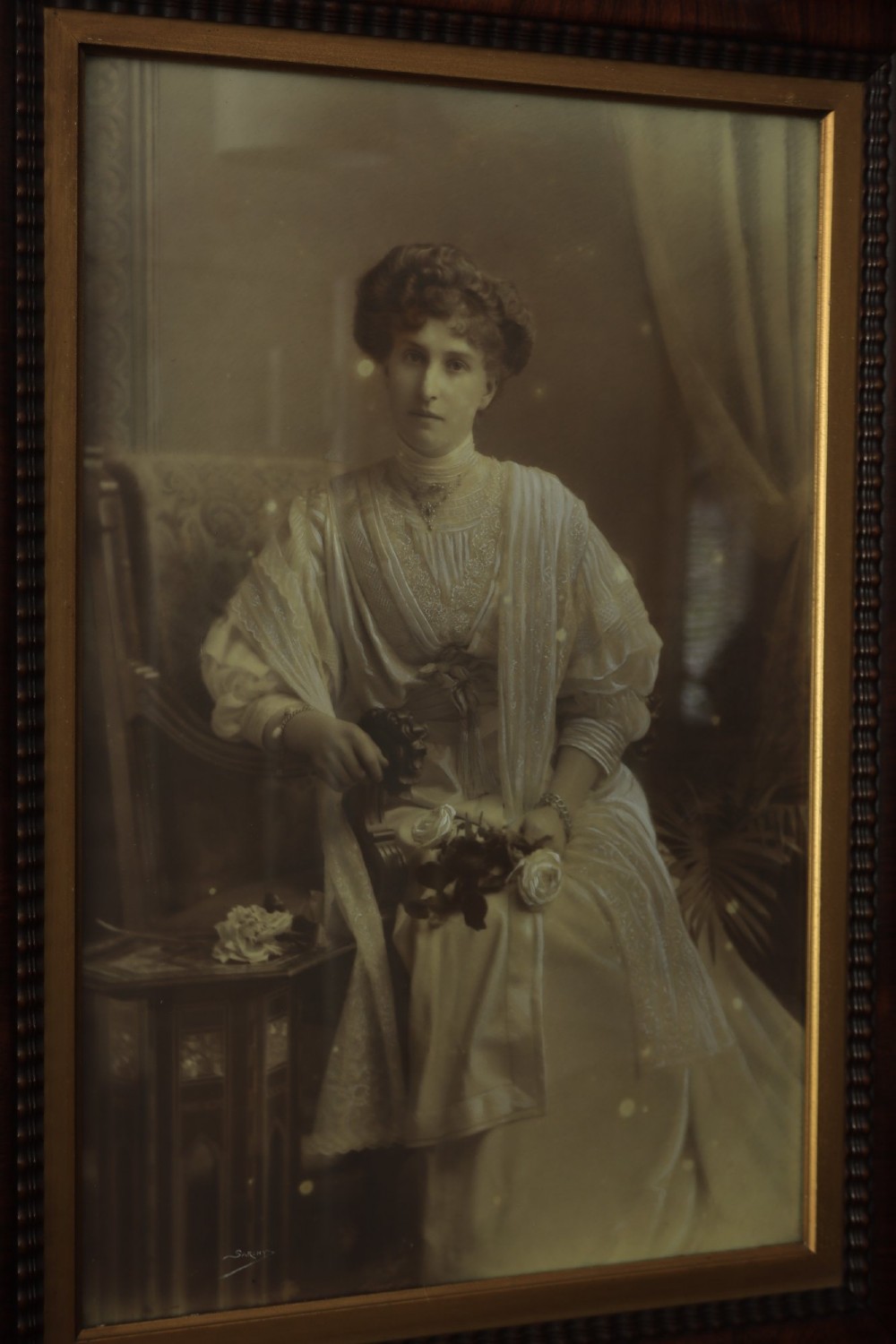 A monochrome Edwardian studio portrait photograph of a seated lady, mounted within a dark polished mahogany frame featuring a gilded inner slip. View VII