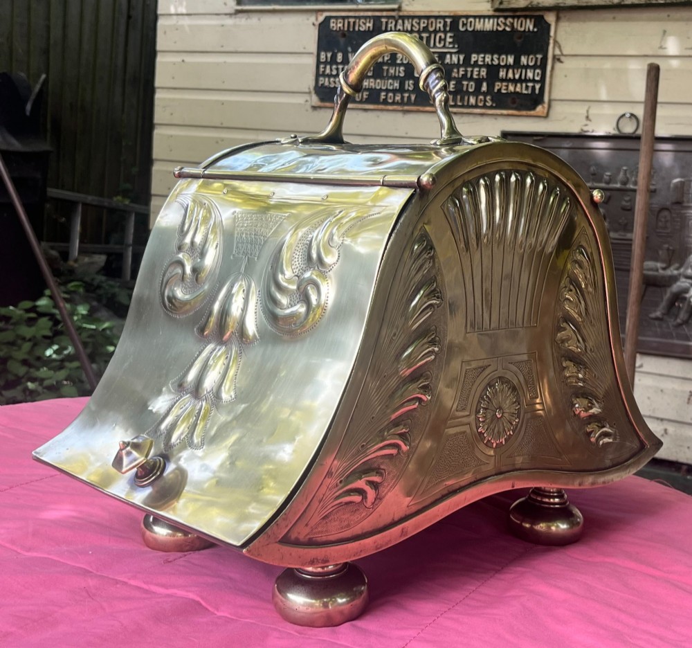 An embossed brass coal scuttle featuring a sloping hinged lid, raised foliate repousse patterns, an integrated top handle, and four raised bun feet.