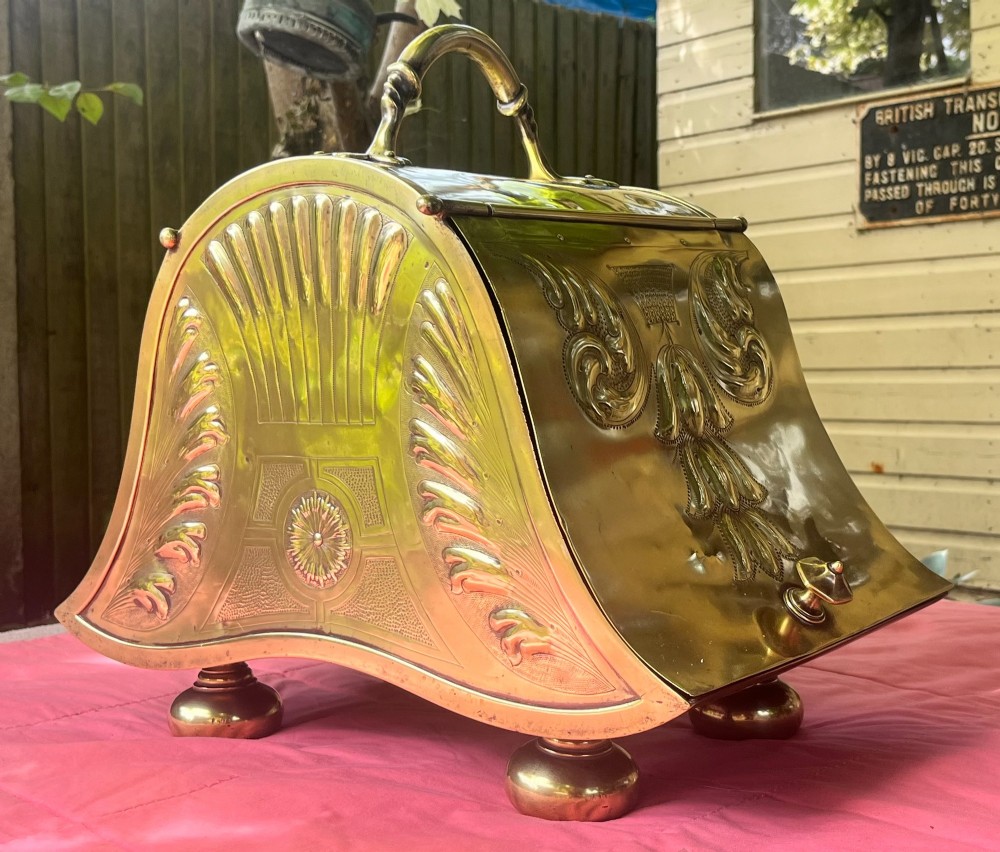 An embossed brass coal scuttle featuring a sloping hinged lid, raised foliate repousse patterns, an integrated top handle, and four raised bun feet. View II