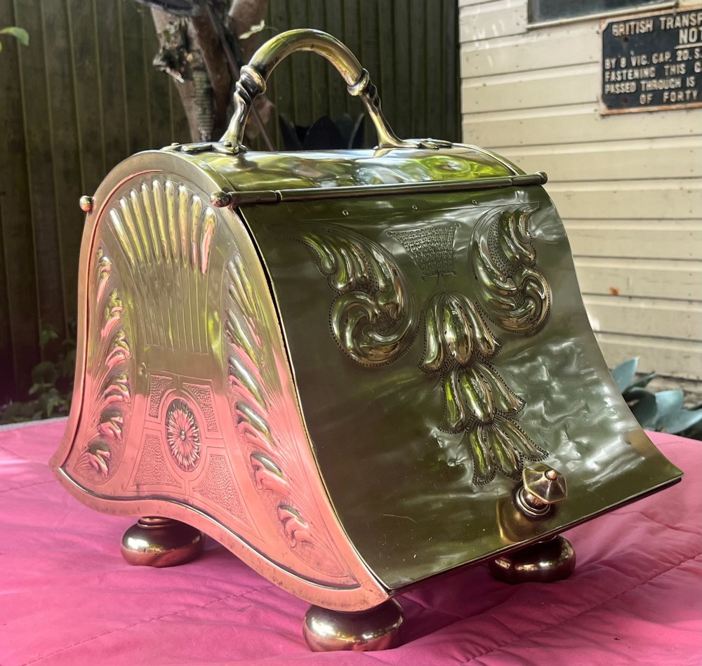 An embossed brass coal scuttle featuring a sloping hinged lid, raised foliate repousse patterns, an integrated top handle, and four raised bun feet. View III