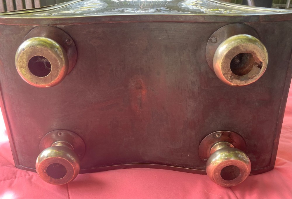 An embossed brass coal scuttle featuring a sloping hinged lid, raised foliate repousse patterns, an integrated top handle, and four raised bun feet. View V