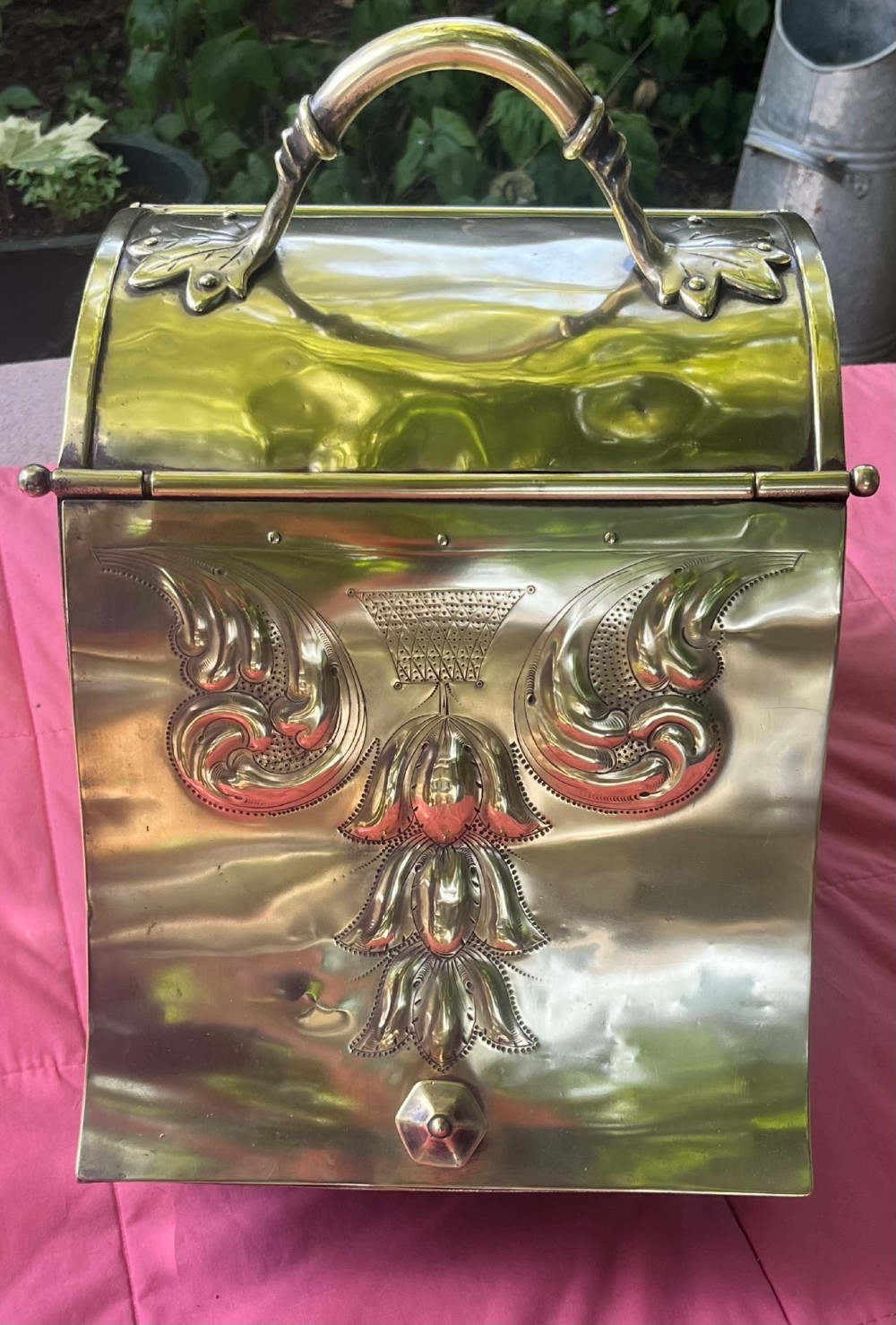 An embossed brass coal scuttle featuring a sloping hinged lid, raised foliate repousse patterns, an integrated top handle, and four raised bun feet. View VII