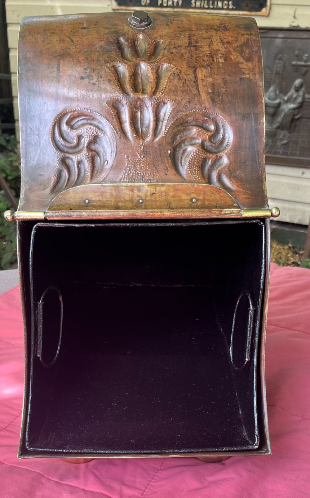 An embossed brass coal scuttle featuring a sloping hinged lid, raised foliate repousse patterns, an integrated top handle, and four raised bun feet. View X
