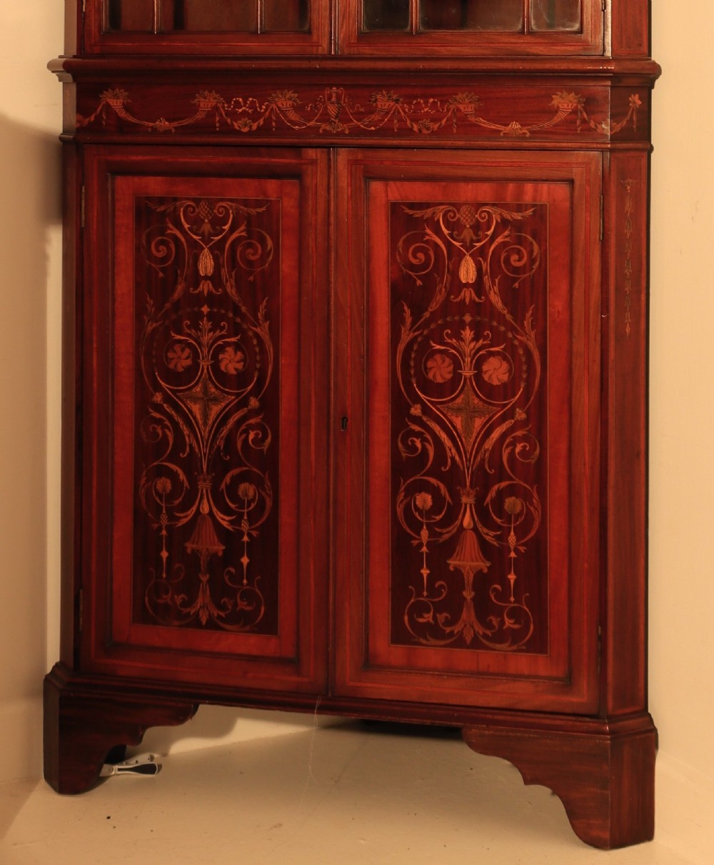 A tall mahogany standing corner cupboard featuring a broken pediment, upper glass doors with Gothic astragal glazing bars, and lower panel doors detailed with intricate marquetry inlay. View II
