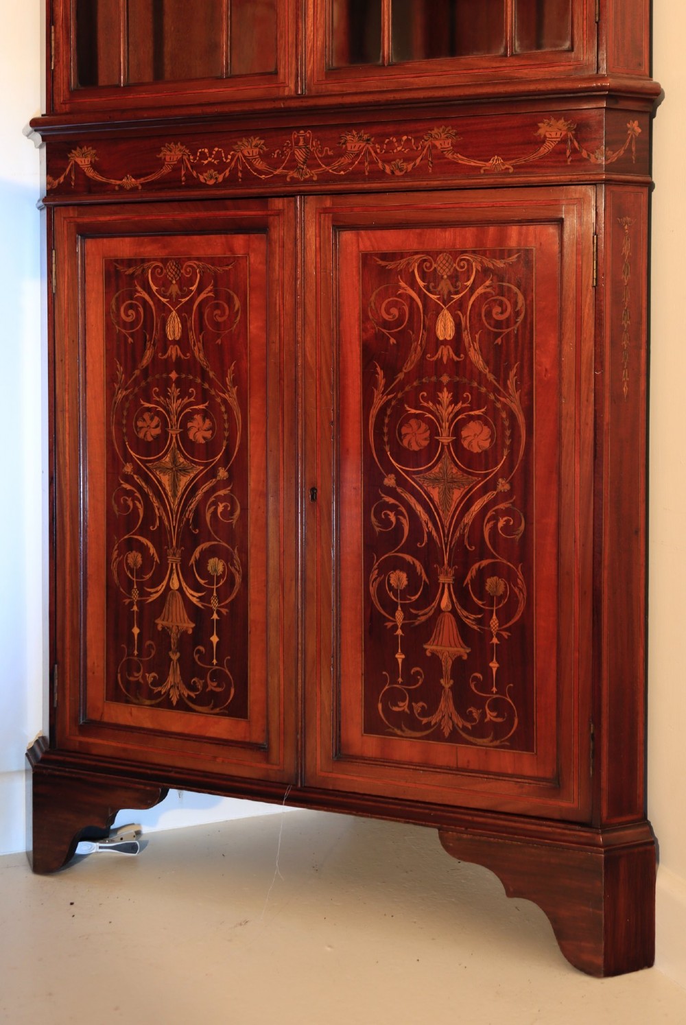 A tall mahogany standing corner cupboard featuring a broken pediment, upper glass doors with Gothic astragal glazing bars, and lower panel doors detailed with intricate marquetry inlay. View IV