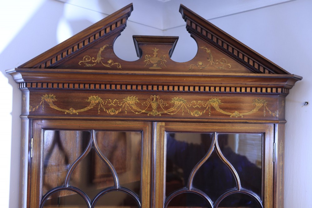 A tall mahogany standing corner cupboard featuring a broken pediment, upper glass doors with Gothic astragal glazing bars, and lower panel doors detailed with intricate marquetry inlay. View VI