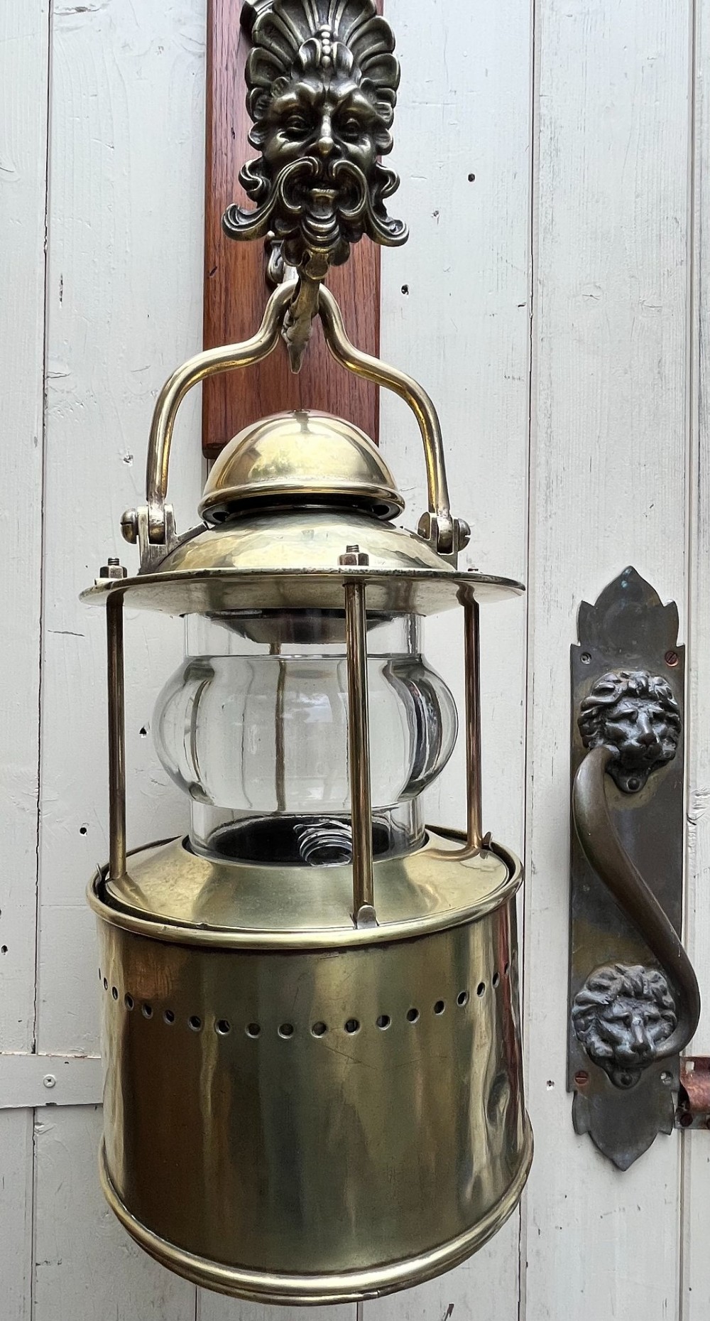 An antique brass wall lantern featuring a clear glass globe enclosed in a vented brass frame, suspended from a cast brass mascaron wall mount.