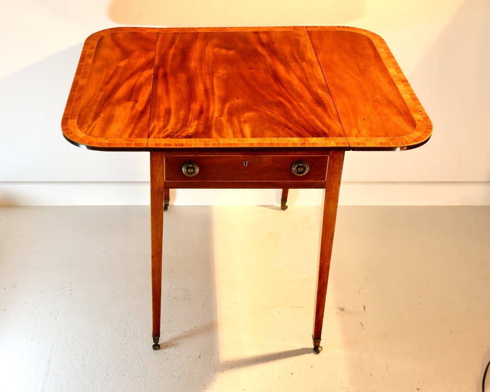 An antique mahogany Pembroke table featuring drop-leaf sides, crossbanded inlay borders, a single drawer with brass handles, and tapered square legs on brass castors.