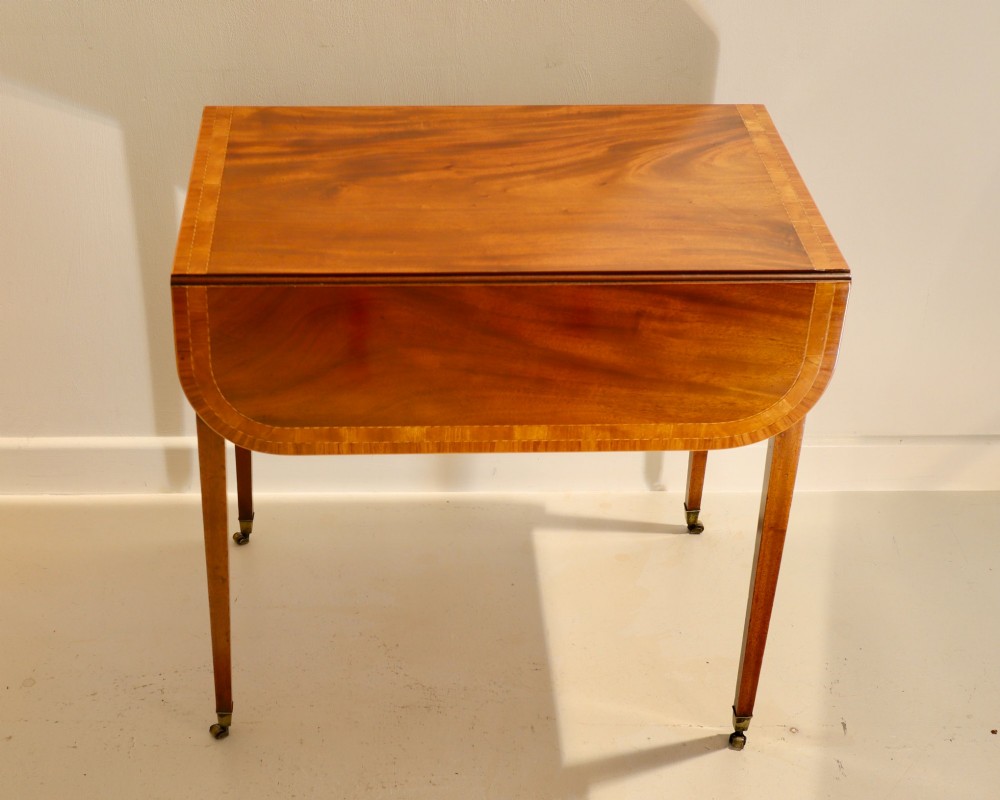 An antique mahogany Pembroke table featuring drop-leaf sides, crossbanded inlay borders, a single drawer with brass handles, and tapered square legs on brass castors. View II