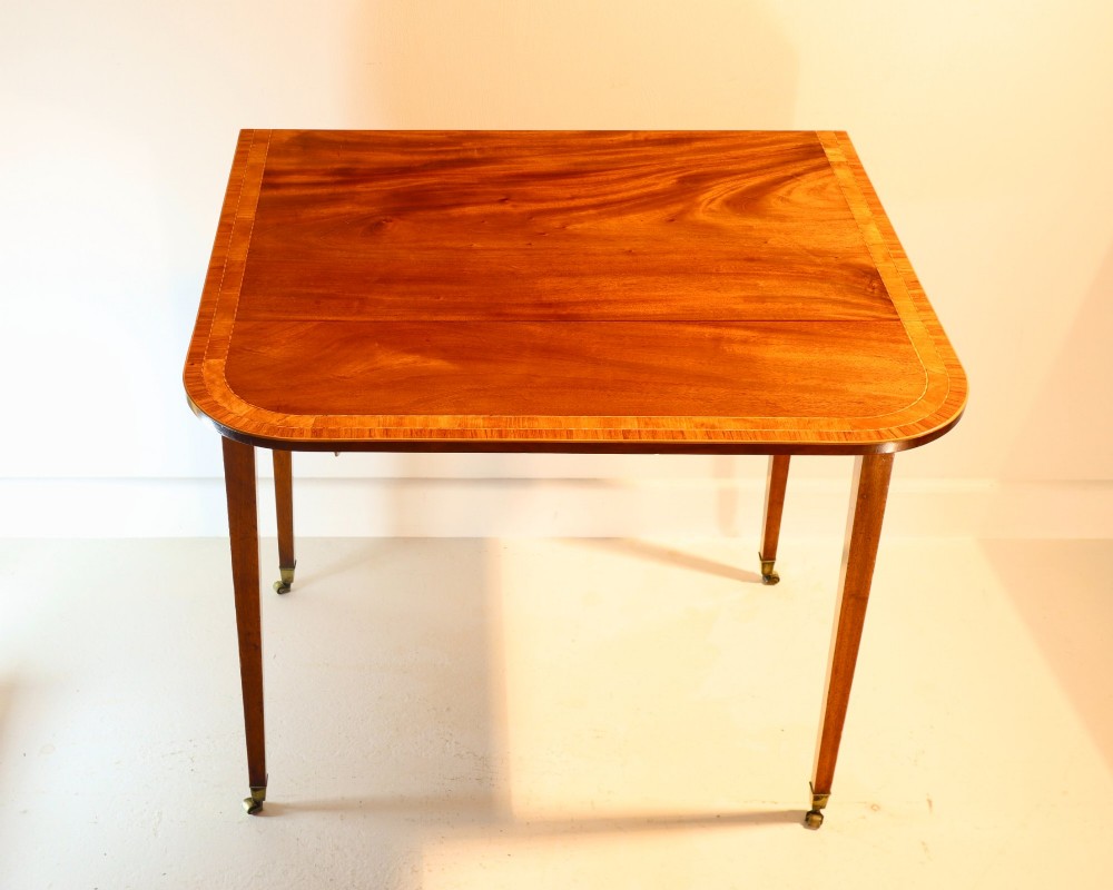 An antique mahogany Pembroke table featuring drop-leaf sides, crossbanded inlay borders, a single drawer with brass handles, and tapered square legs on brass castors. View III