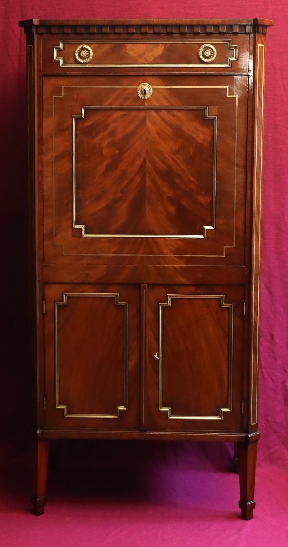 An antique mahogany escritoire featuring brass string inlay, a top drawer, a fall-front writing panel, and a two-door lower cupboard raised on tapered legs.