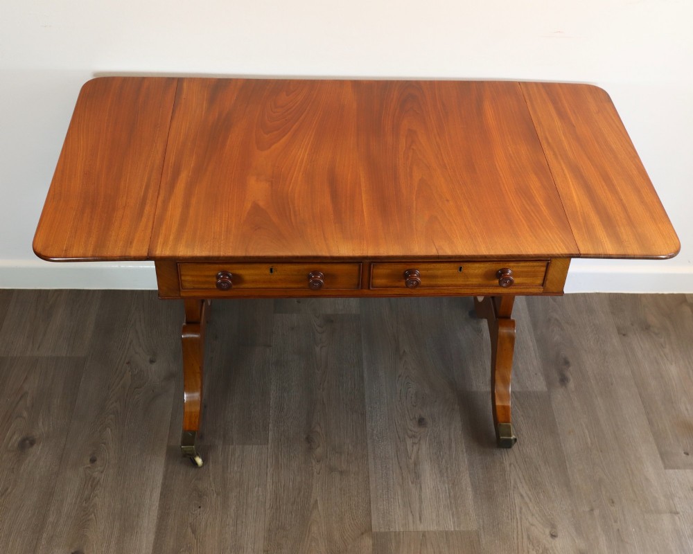 A Regency mahogany sofa table featuring two drop-leaf ends, two frieze drawers with turned wooden handles, and splayed legs with brass castors.