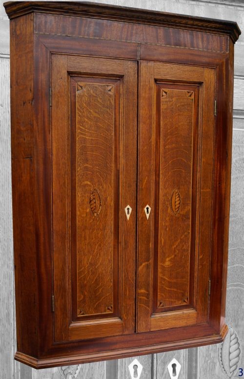 An antique oak hanging corner cupboard with twin panelled doors, inlaid central motifs, bone keyhole escutcheons, and a moulded top cornice.
