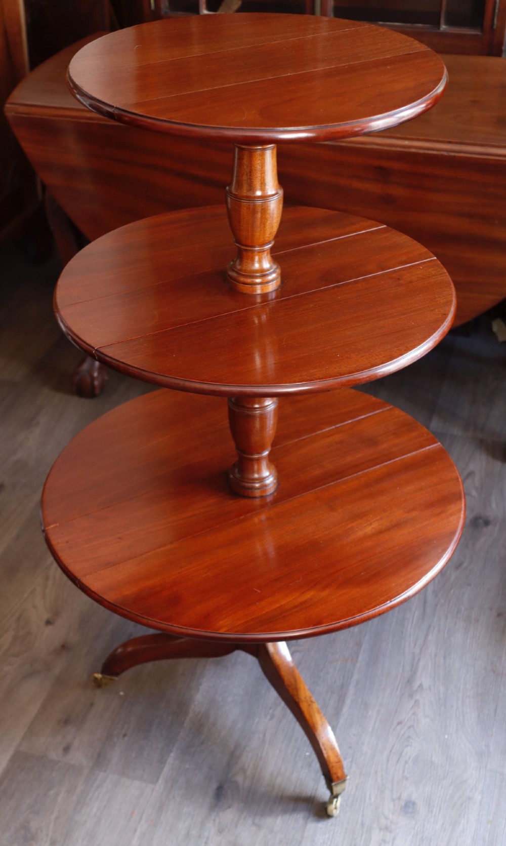 A three-tiered mahogany dumb waiter featuring circular graduating surfaces supported by turned central columns and a tripod base on brass castors.