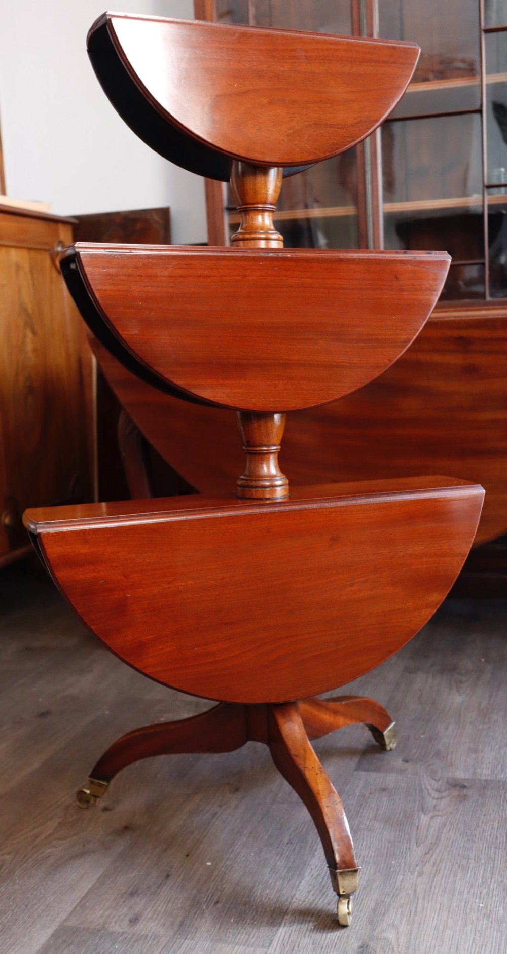 A three-tiered mahogany dumb waiter featuring circular graduating surfaces supported by turned central columns and a tripod base on brass castors. View II