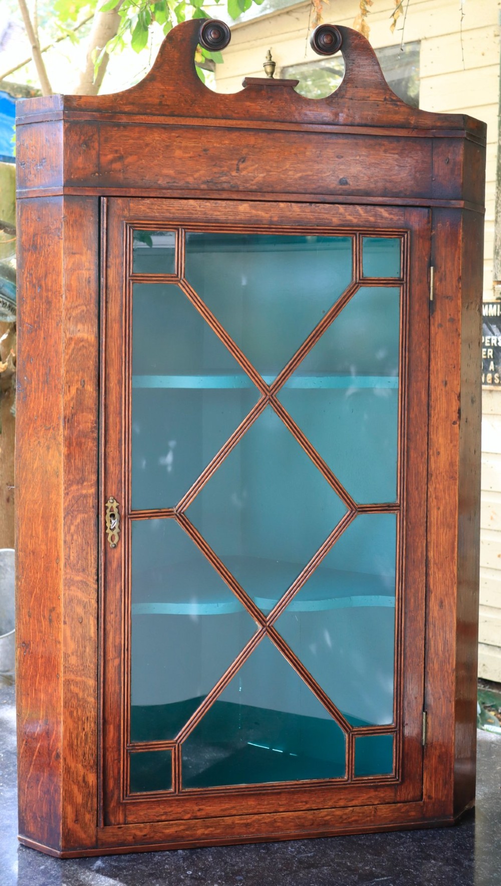 An antique wooden hanging corner cabinet featuring an astragal glazed door, a swan-neck pediment, and a painted teal interior with two shelves.