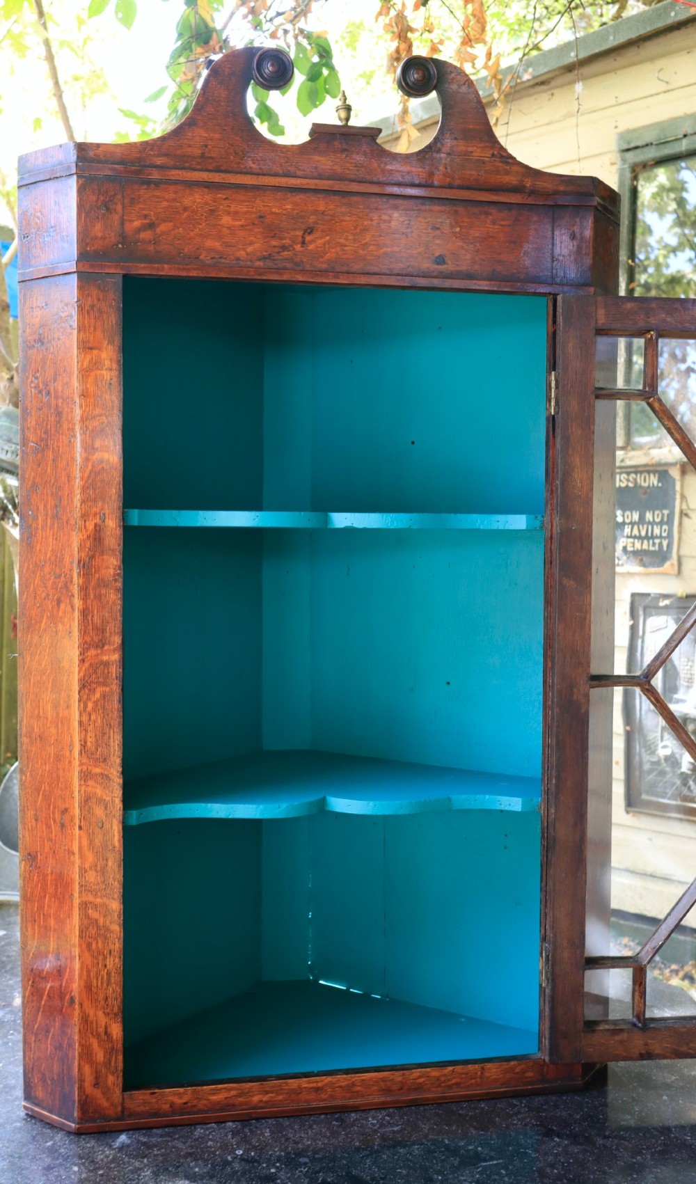 An antique wooden hanging corner cabinet featuring an astragal glazed door, a swan-neck pediment, and a painted teal interior with two shelves. View II