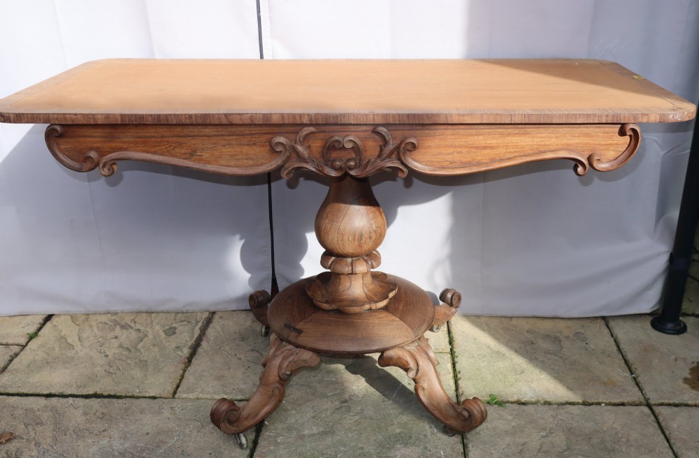 A Victorian rosewood centre table with a rectangular top, carved frieze, a central turned bulbous pedestal, and scroll legs on castors.