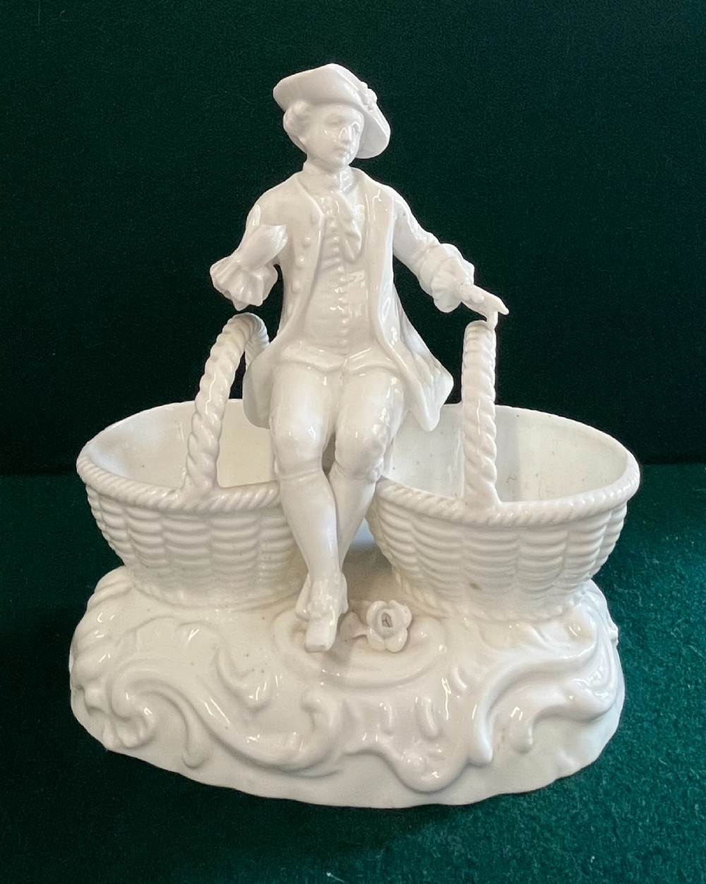 A pair of white glazed porcelain figurines depicting a male and female figure in 18th-century attire, each seated between two woven basket-form dishes on scrollwork bases. View II