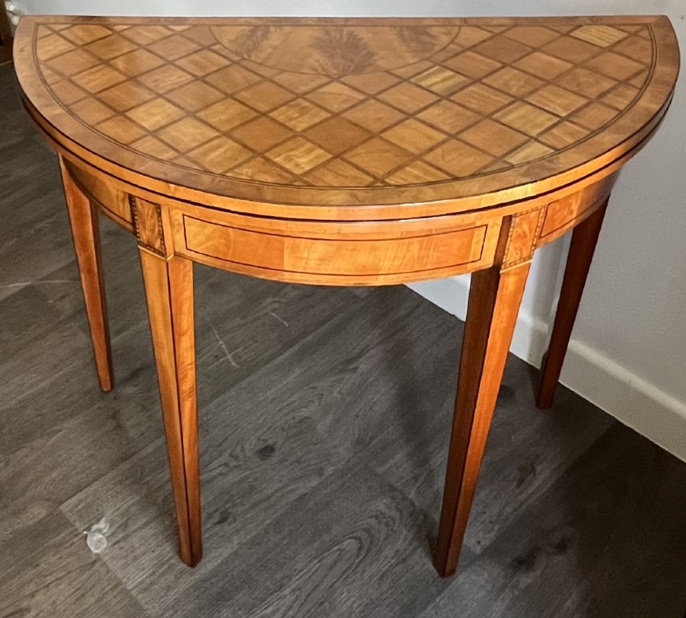 An antique George III demilune card table in honey satinwood featuring geometric parquetry inlay and tapered legs.