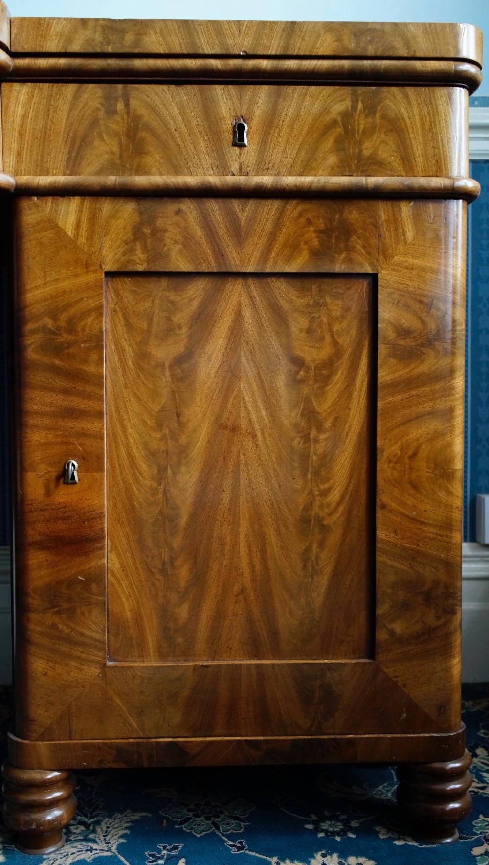 A flame mahogany pedestal desk with a central green leather writing surface, open upper shelf gallery, and turned feet. View III