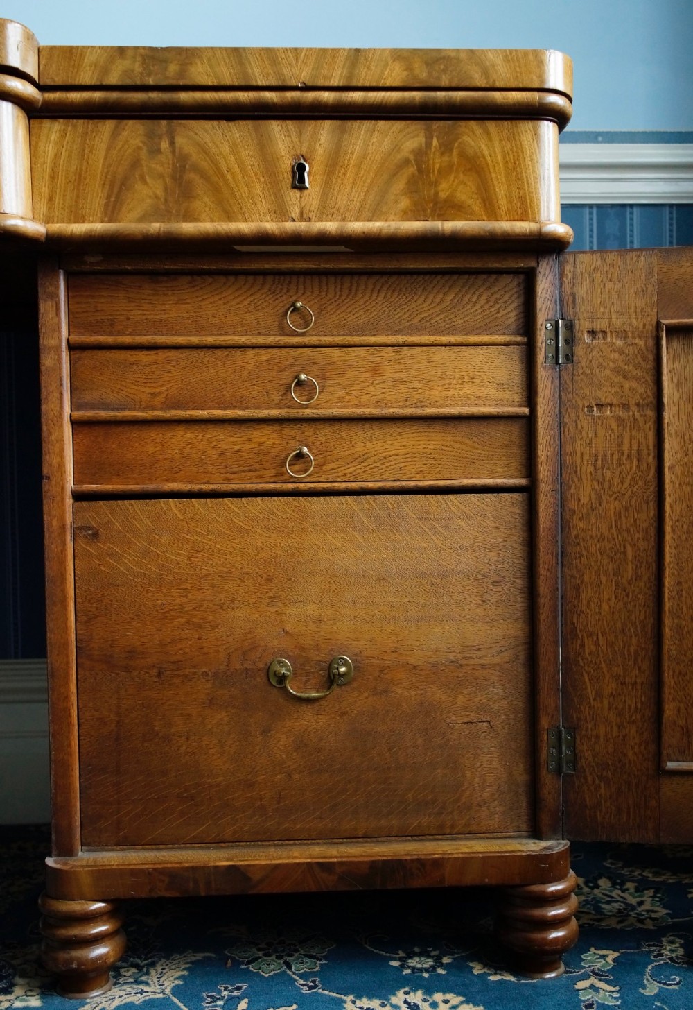 A flame mahogany pedestal desk with a central green leather writing surface, open upper shelf gallery, and turned feet. View IV
