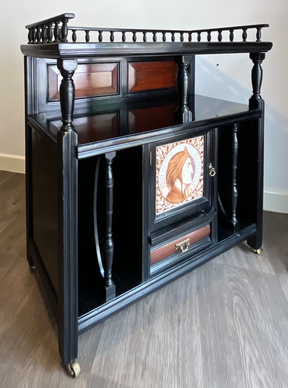 A late Victorian ebonised wood cabinet with a top spindle gallery, dark mahogany panels, and a central door featuring a profile ceramic tile of Dante. View II
