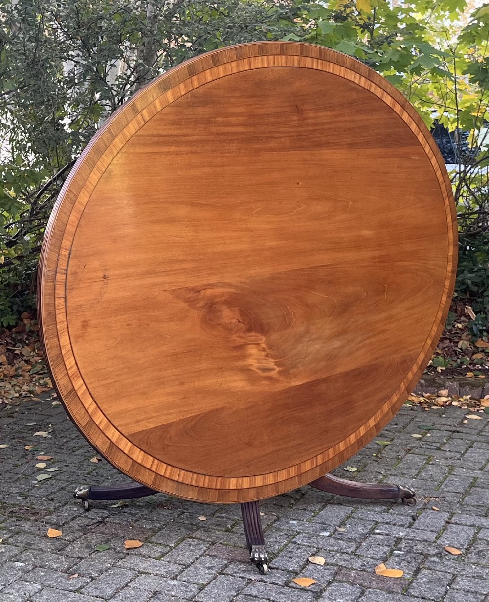 An antique wood tilt-top dining table displaying a circular crossbanded top with inlaid border on a central pedestal base.