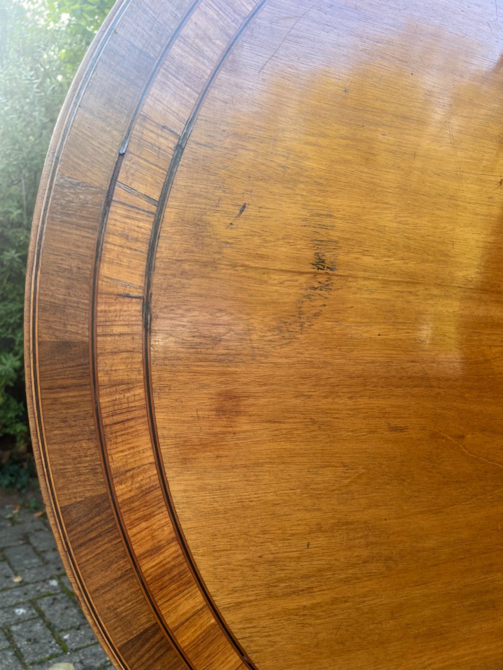 An antique wood tilt-top dining table displaying a circular crossbanded top with inlaid border on a central pedestal base. View III
