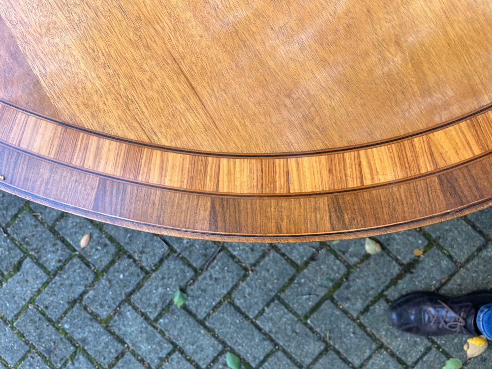 An antique wood tilt-top dining table displaying a circular crossbanded top with inlaid border on a central pedestal base. View VI
