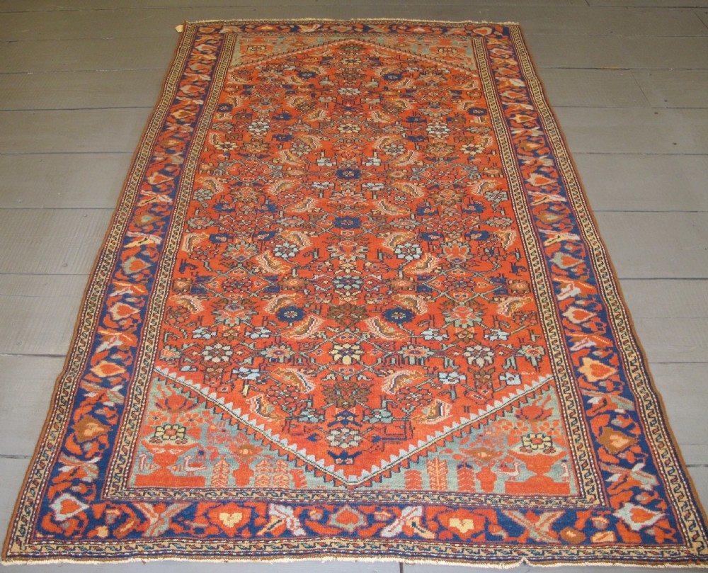 An antique hand-knotted West Persian wool rug featuring a central rust red field with geometric motifs and a dark blue patterned border.