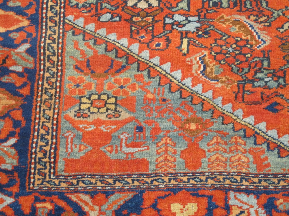 An antique hand-knotted West Persian wool rug featuring a central rust red field with geometric motifs and a dark blue patterned border. View II