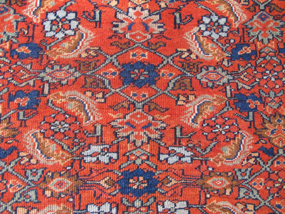 An antique hand-knotted West Persian wool rug featuring a central rust red field with geometric motifs and a dark blue patterned border. View III