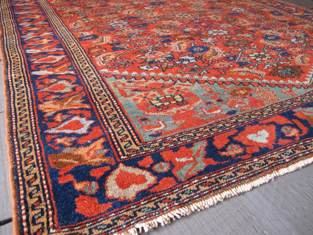An antique hand-knotted West Persian wool rug featuring a central rust red field with geometric motifs and a dark blue patterned border. View IV