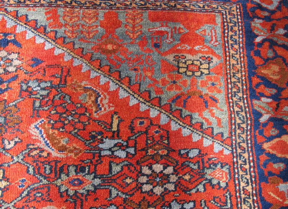 An antique hand-knotted West Persian wool rug featuring a central rust red field with geometric motifs and a dark blue patterned border. View V