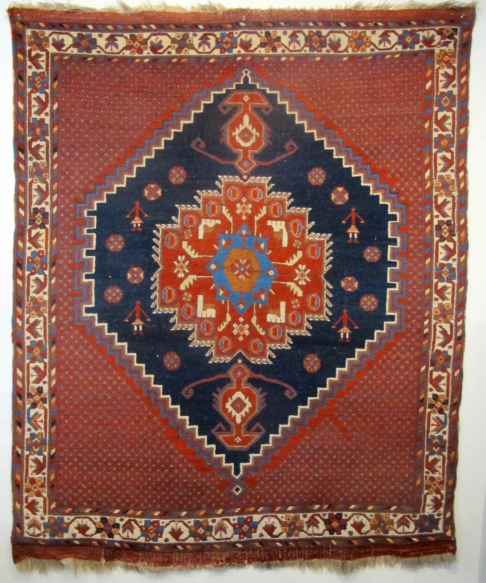 An antique Persian tribal rug featuring a central navy blue stepped medallion on a dark red geometric field, framed by stylized decorative borders.