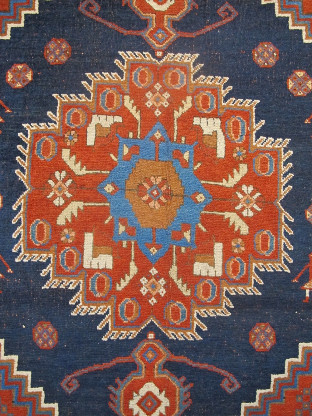 An antique Persian tribal rug featuring a central navy blue stepped medallion on a dark red geometric field, framed by stylized decorative borders. View III