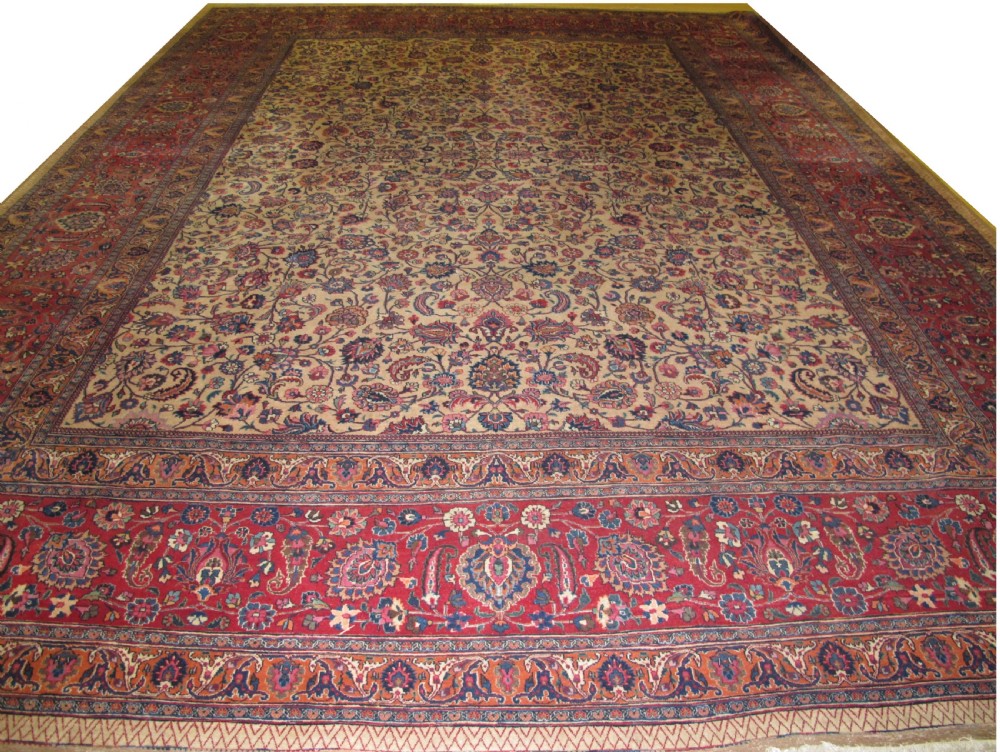 A large antique Persian wool carpet featuring a dense all-over floral pattern on a cream field framed by a wide red decorative border.